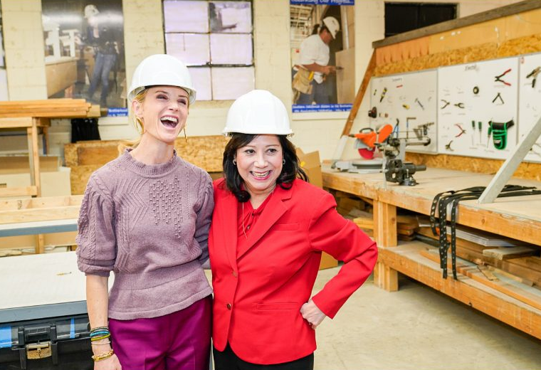 Wearing hard hats, First Partner Jennifer Siebel Newsom and a woman stand arm in arm in front of a workbench.