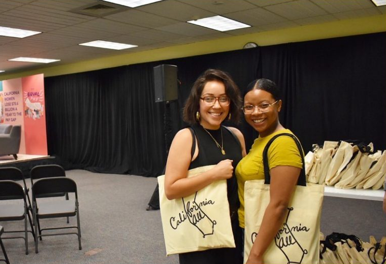 Behind the scenes of the CA Equal Pay conference, two women of color pose with California for All tote bags.