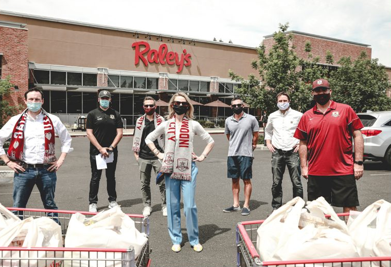he First Partner, a blonde, light-skinned woman, stands with hands on her hips in front of six men in the parking lot of a Raley's grocery store. In front, there are two grocery carts filled with plastic shopping bags of food. Everyone is wearing a face mask.